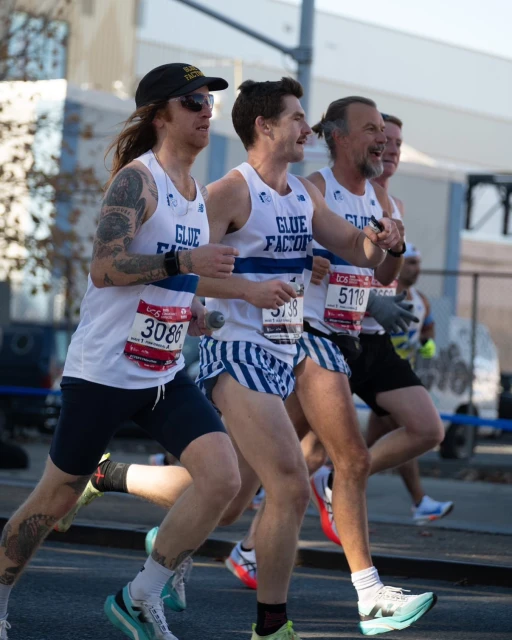 A group of male runners in athletic wear compete in an outdoor road race, wearing numbered bibs and running shoes on a sunny day.