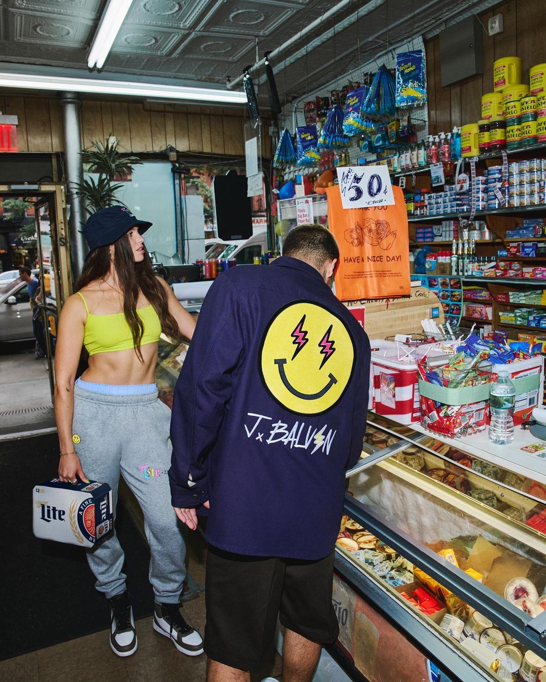 Woman holding beer stands in a bodega next to a man wearing a smiley face jacket, with grocery items displayed in the background.