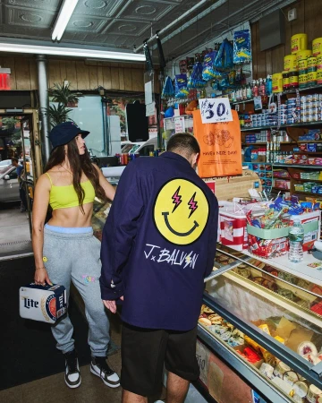 Woman holding beer stands in a bodega next to a man wearing a smiley face jacket, with grocery items displayed in the background.
