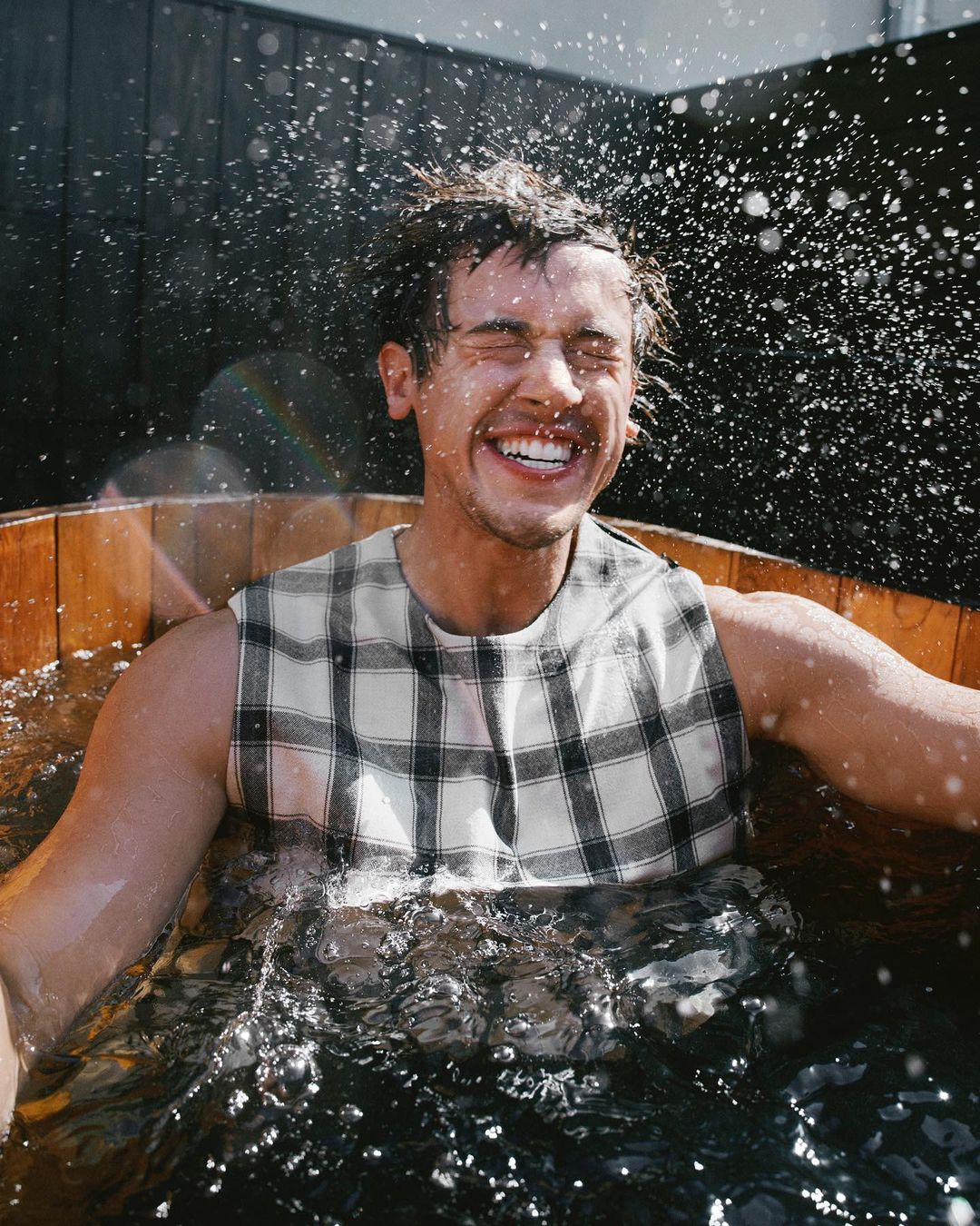 Smiling man in a wooden barrel tub with splashing water. Wearing a sleeveless plaid shirt, eyes closed, teeth showing. Outdoor setting.
