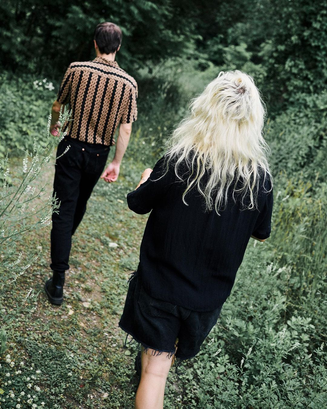 Two people walk a grassy path lined with green foliage. One person wears a patterned shirt and dark pants, while the other wears all black.