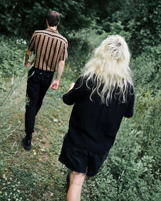 Two people walk a grassy path lined with green foliage. One person wears a patterned shirt and dark pants, while the other wears all black.