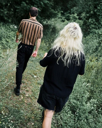 Two people walk a grassy path lined with green foliage. One person wears a patterned shirt and dark pants, while the other wears all black.