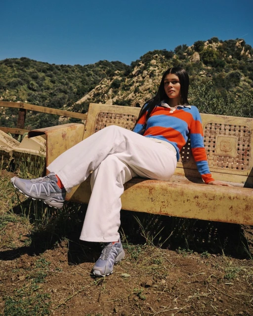 Young woman in sporty casual attire sits on an old bench against a mountain backdrop, wearing trail running shoes and a colorful sweater.