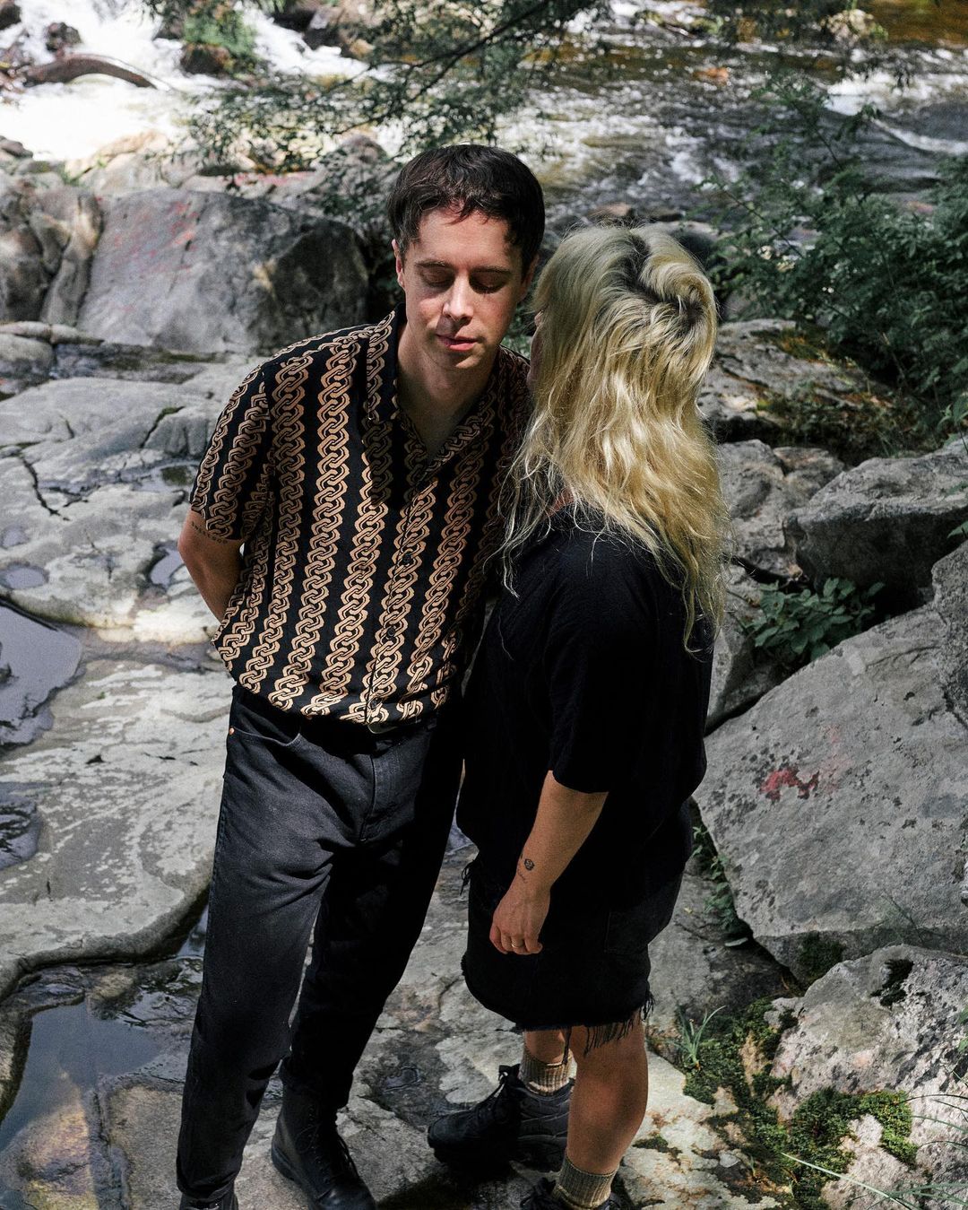 A stylish couple poses in front of a rocky waterfall; the man wears a patterned shirt, and the woman has blonde hair and a black outfit.