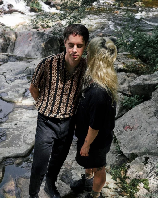 A stylish couple poses in front of a rocky waterfall; the man wears a patterned shirt, and the woman has blonde hair and a black outfit.