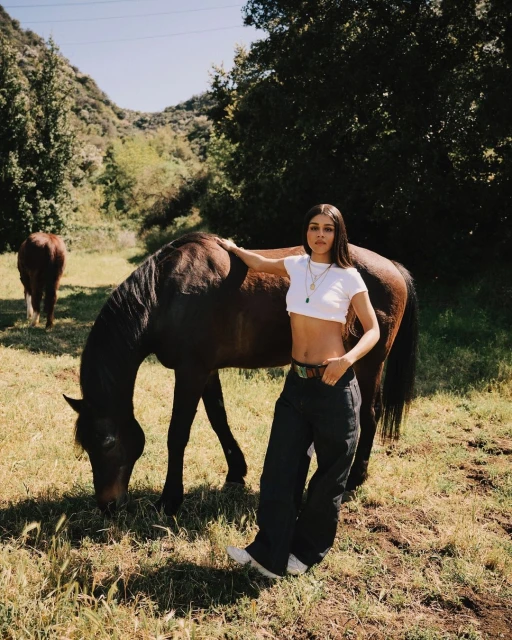 Woman poses with a brown horse in a sunlit meadow with a mountain backdrop, wearing a white crop top and dark pants.