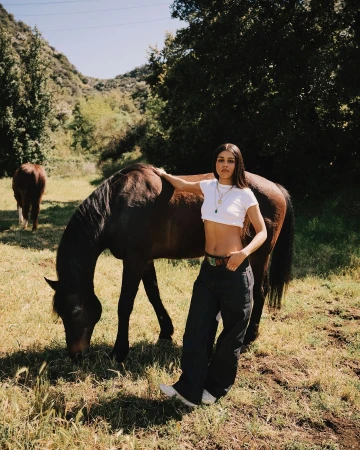 Woman poses with a brown horse in a sunlit meadow with a mountain backdrop, wearing a white crop top and dark pants.
