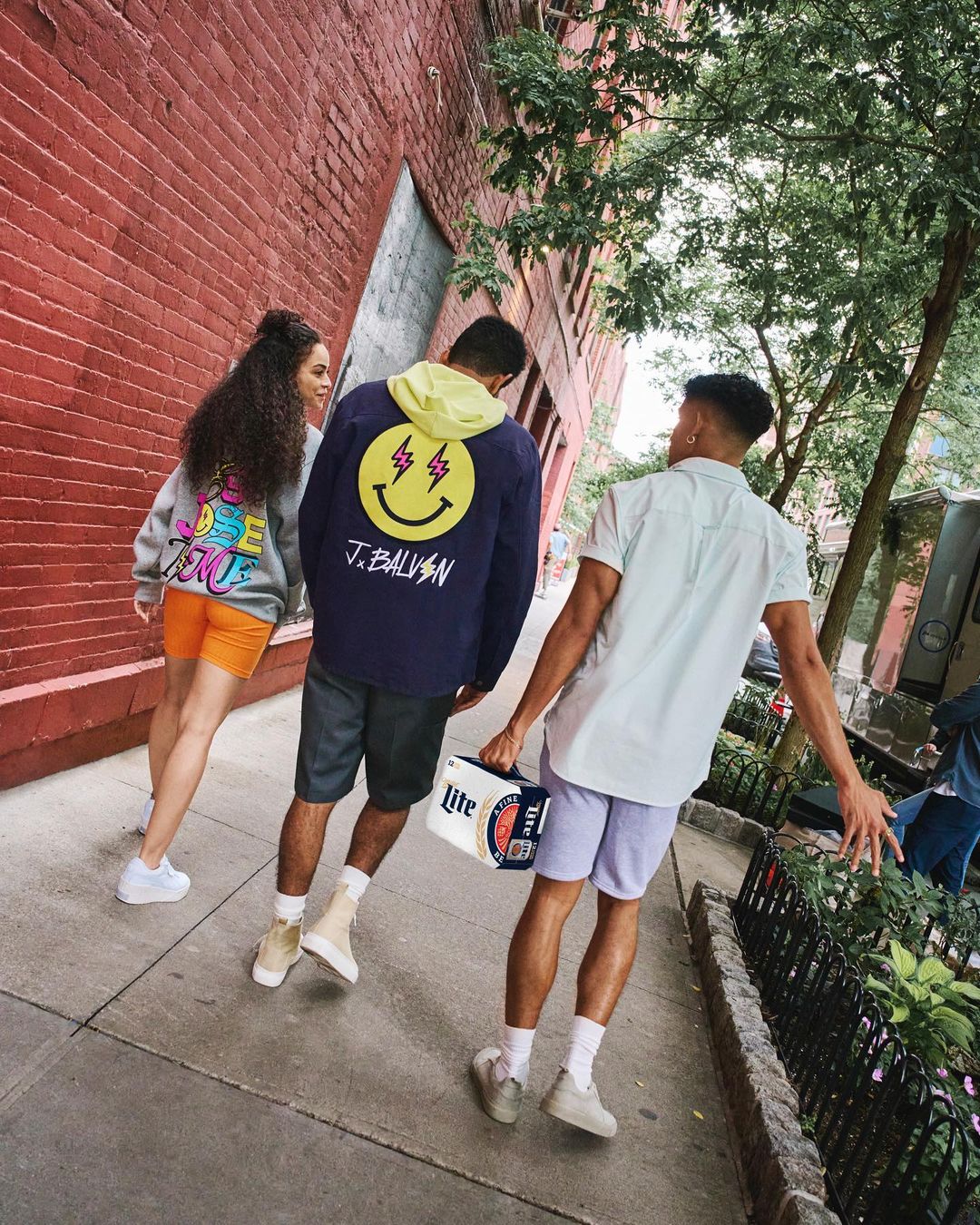 Three people walking on a city sidewalk with a brick wall, trees, and buildings in the background on a sunny day in an urban setting.