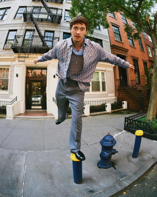 A person in a patterned shirt balancing on a blue pole on a city sidewalk, with apartment buildings and a fire hydrant in the background.