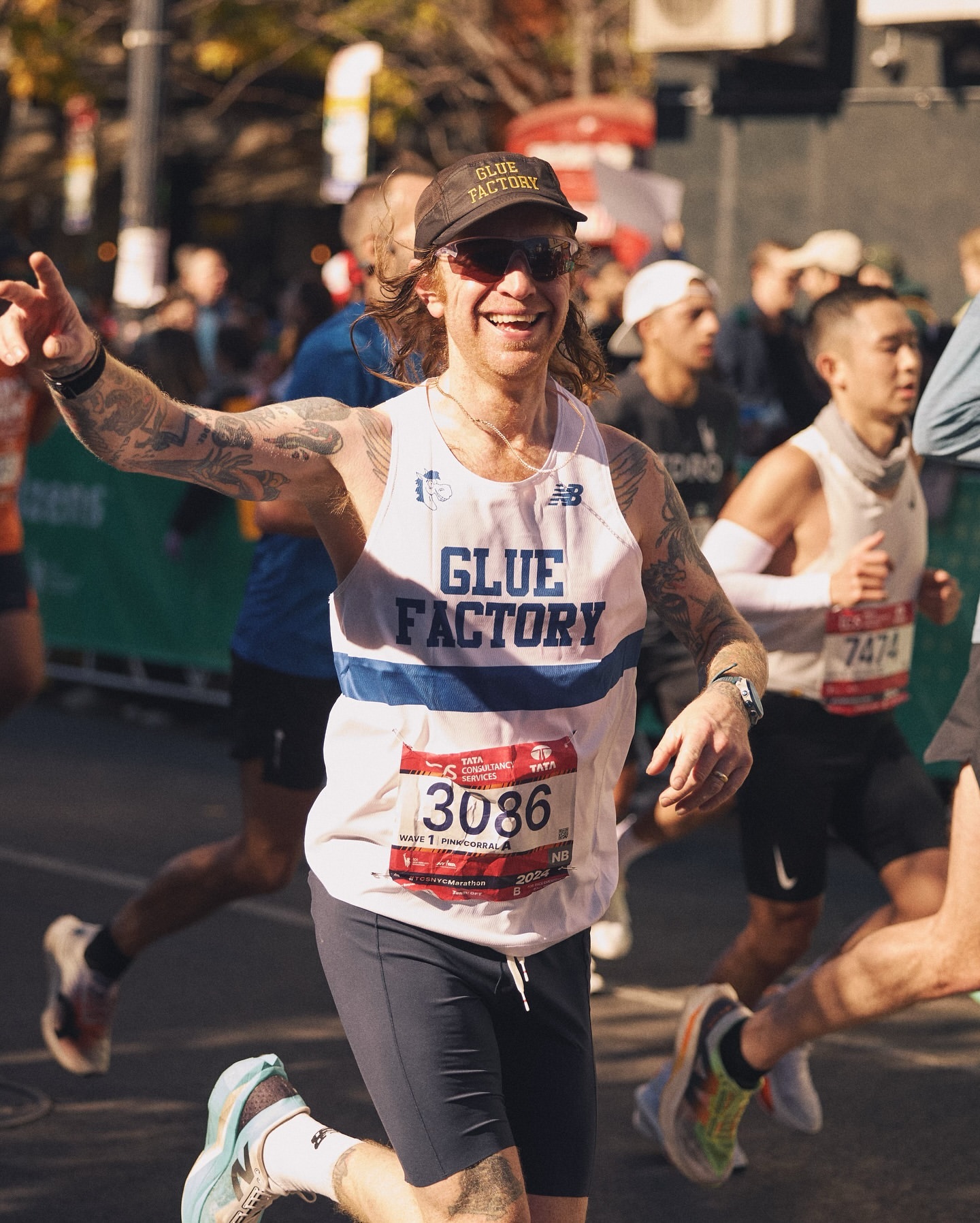 Man smiles broadly while running marathon, wearing a 'Glue Factory' hat and tank top, race number 3086, surrounded by other runners.