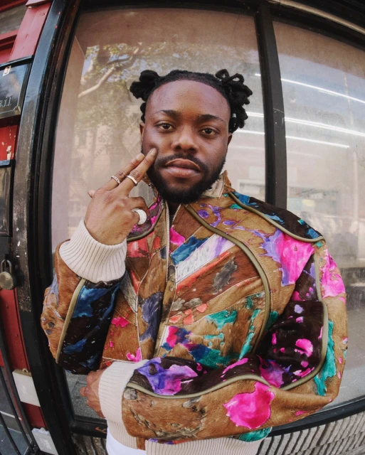 Close-up shot of an adult male with locs, wearing a colorful patchwork jacket with cream-colored ribbed trim, and multiple rings on his fingers.