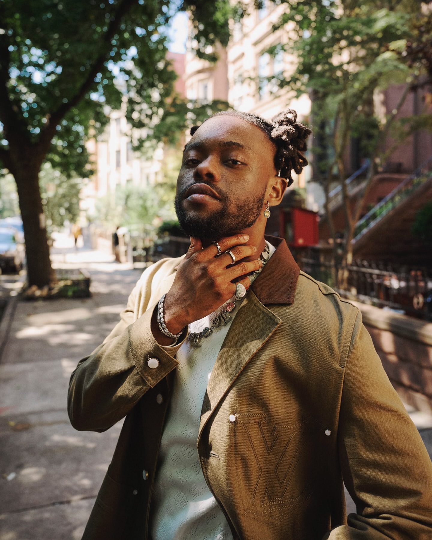 Portrait of an adult male wearing a tan jacket and silver chain, posing outdoors against a background of leafy trees and urban buildings.