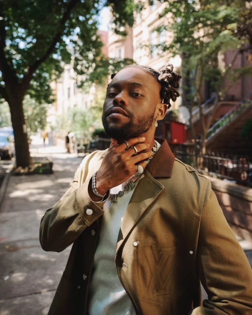 Portrait of an adult male wearing a tan jacket and silver chain, posing outdoors against a background of leafy trees and urban buildings.