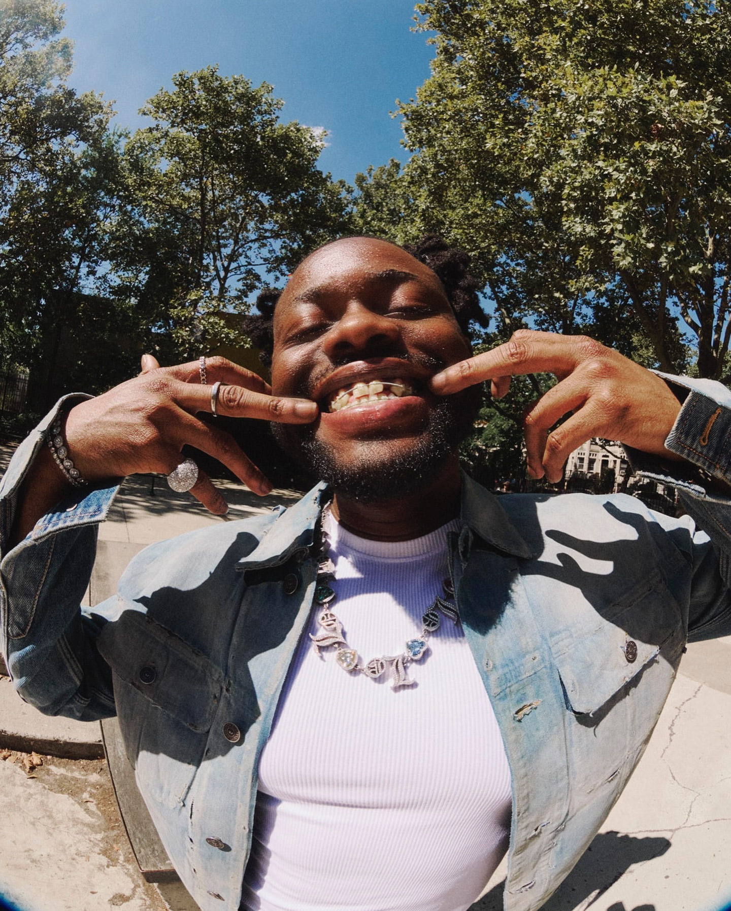 Smiling Black adult male with dark hair wearing jewelry, denim jacket, and white top outdoors with trees and bright blue sky.