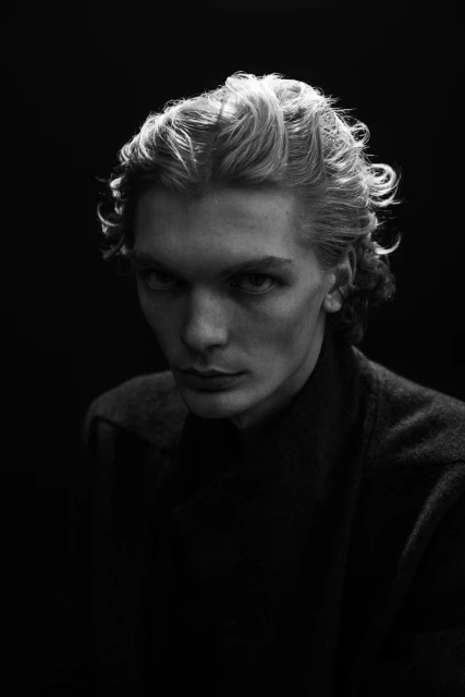 Black and white portrait of a fair-skinned adult man with wavy blonde hair wearing a dark shirt and jacket against a black background.