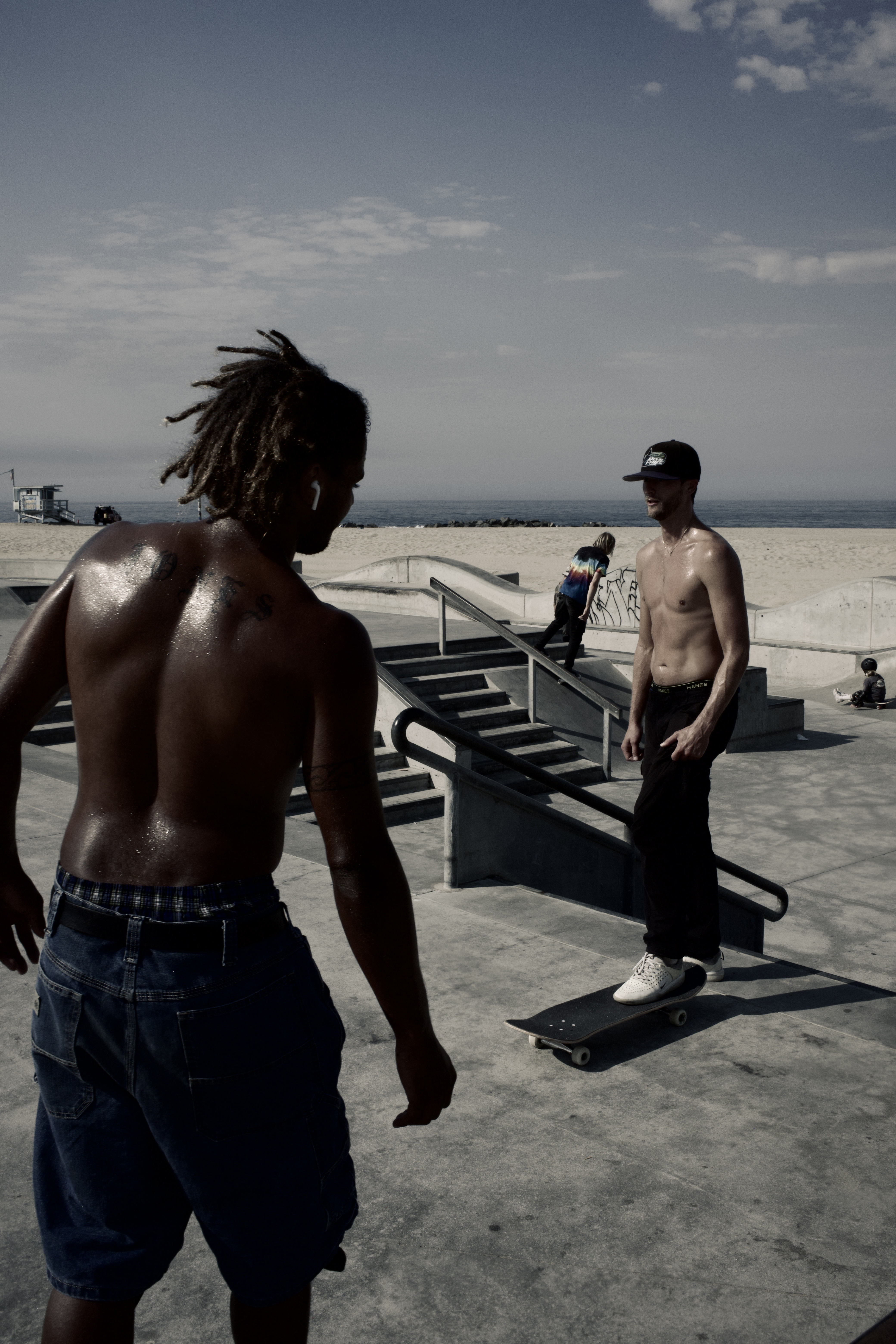 Two shirtless men in an outdoor skatepark on a sunny day, one with dreadlocks facing away, one on a skateboard, blue sky and beach.