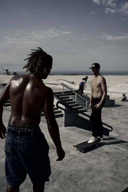 Two shirtless men in an outdoor skatepark on a sunny day, one with dreadlocks facing away, one on a skateboard, blue sky and beach.