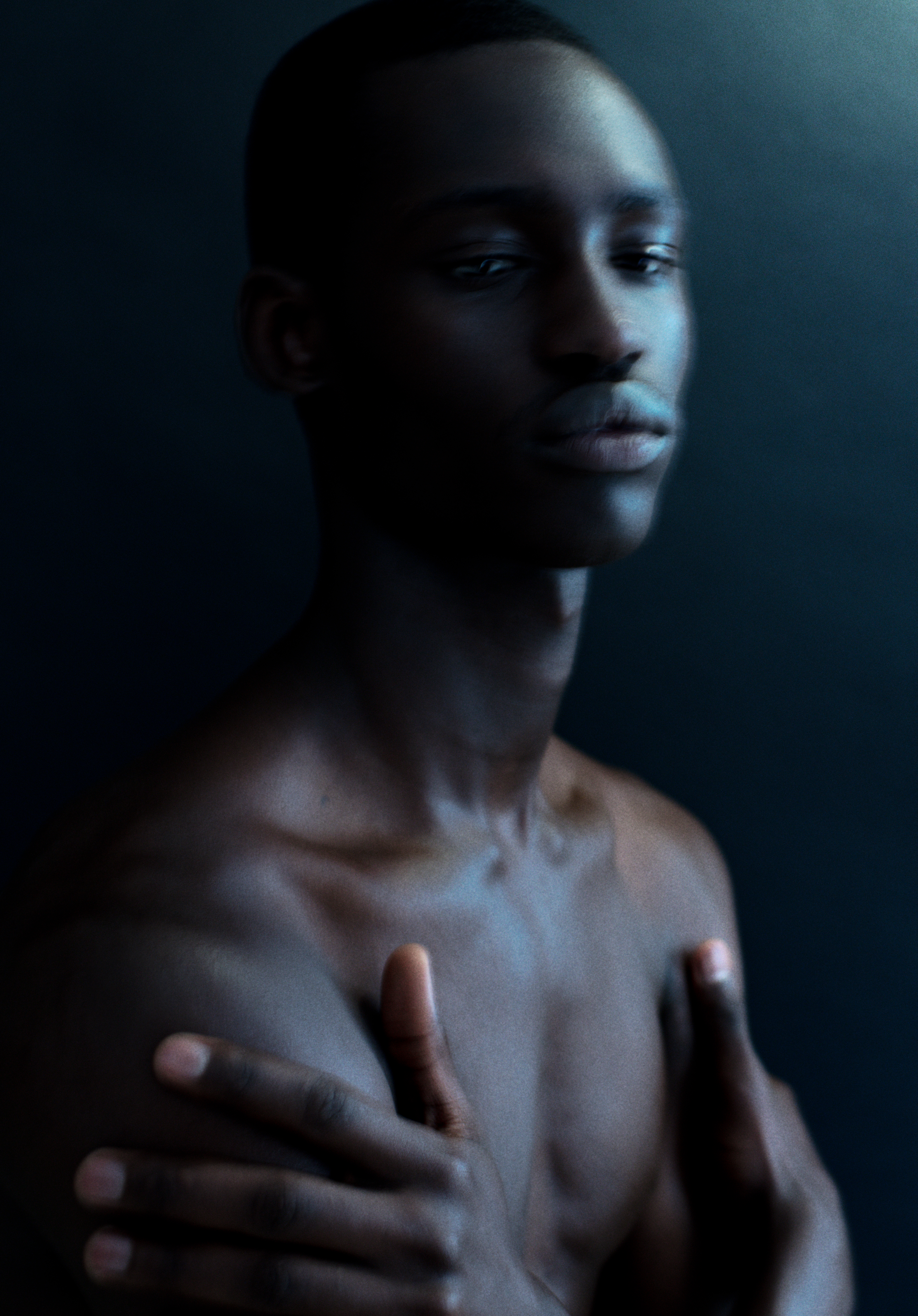 Striking portrait of a shirtless adult male with dark skin, crossed arms, and a calm expression against a dark blue background.