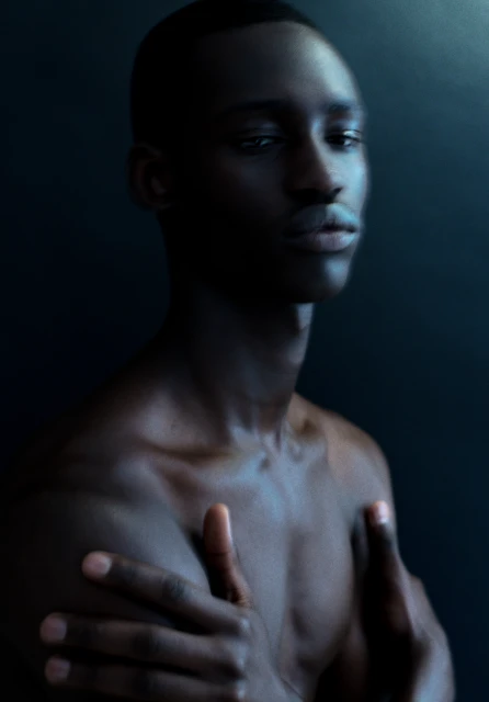 Striking portrait of a shirtless adult male with dark skin, crossed arms, and a calm expression against a dark blue background.