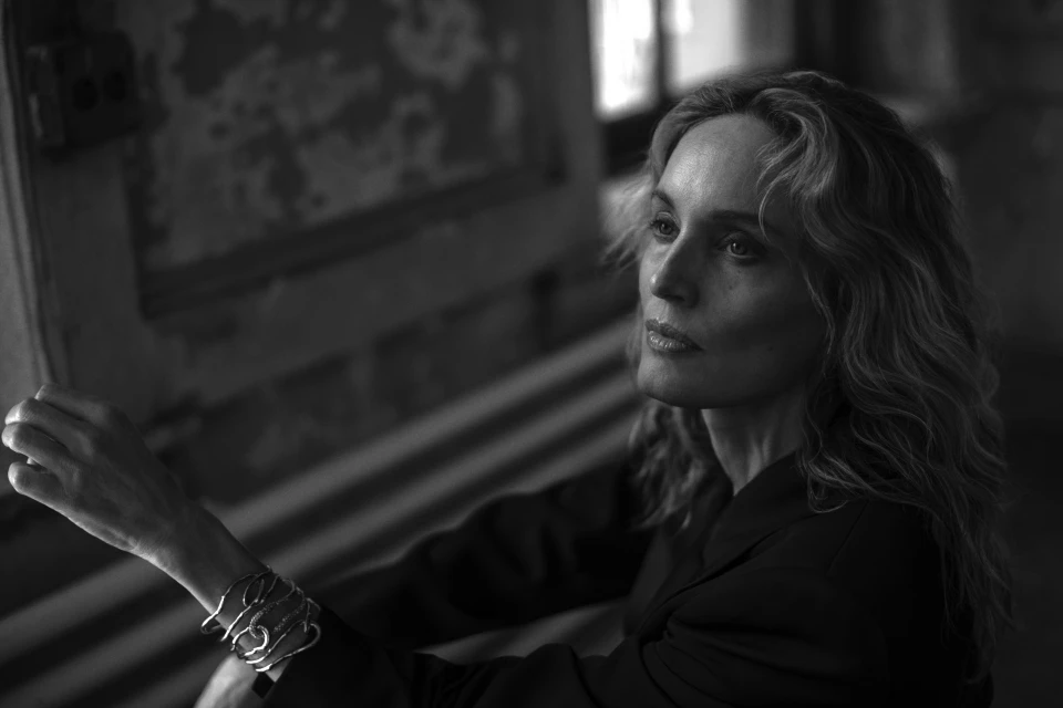 Black and white portrait of an adult woman with wavy blonde hair wearing a blazer and bracelet against a textured wall.
