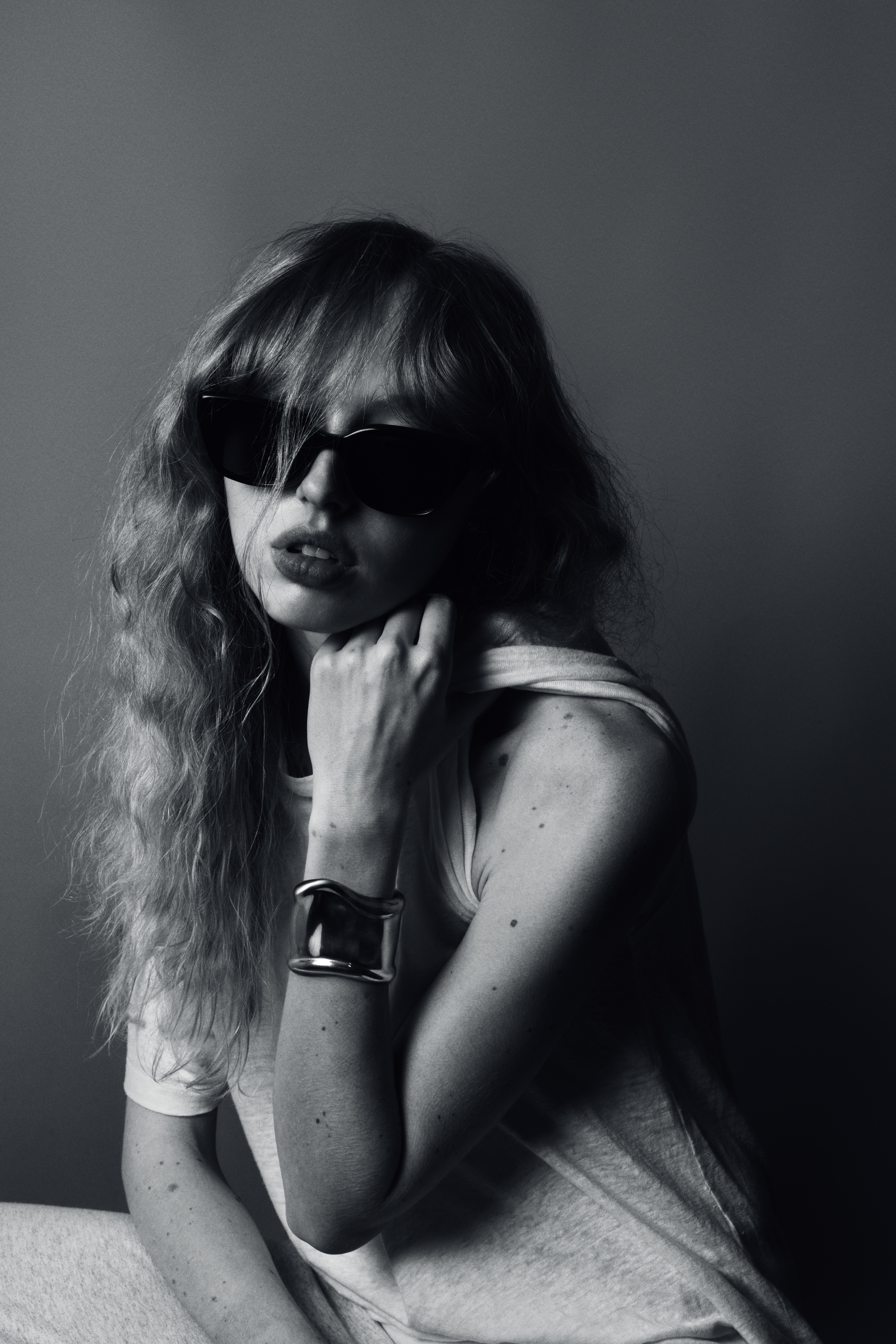 Black and white portrait of a woman with curly hair and sunglasses wearing a cuff bracelet in front of a gray backdrop.