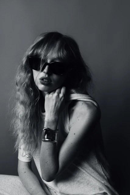 Black and white portrait of a woman with curly hair and sunglasses wearing a cuff bracelet in front of a gray backdrop.