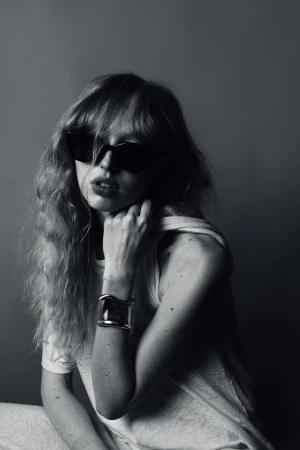 Black and white portrait of a woman with curly hair and sunglasses wearing a cuff bracelet in front of a gray backdrop.