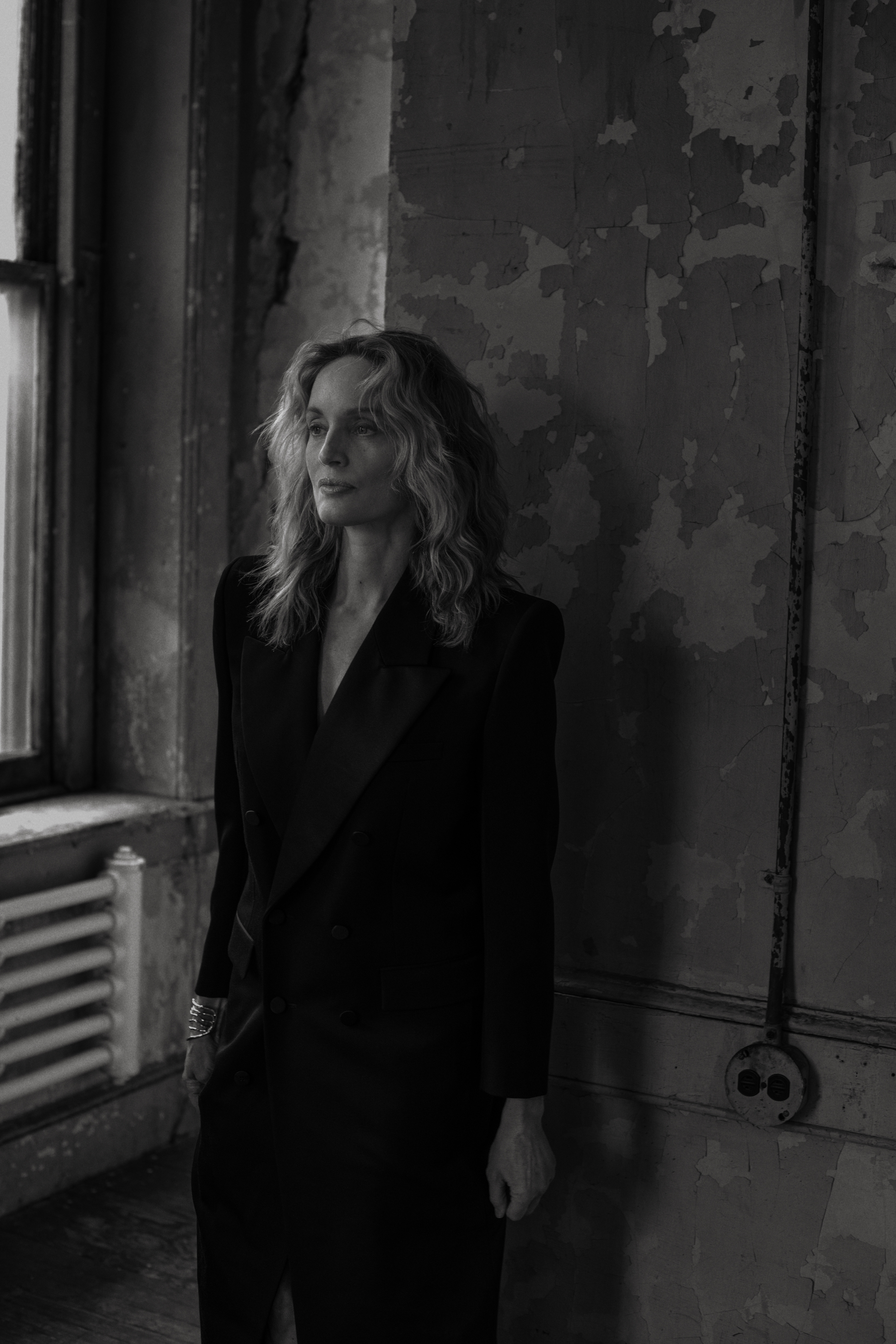 Black and white portrait of a woman in a black dress posing in an old building with peeling paint and natural light from a window.