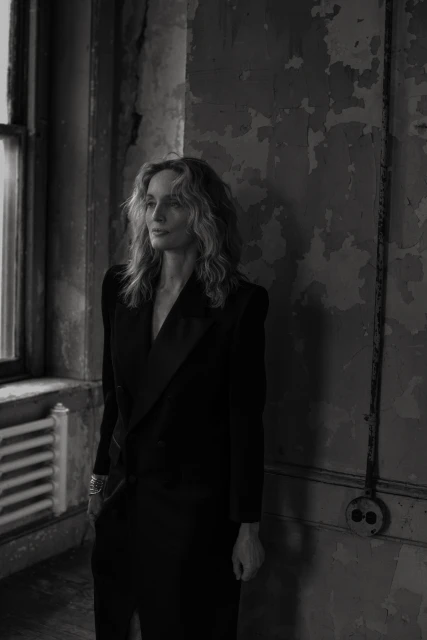 Black and white portrait of a woman in a black dress posing in an old building with peeling paint and natural light from a window.