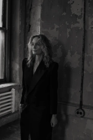 Black and white portrait of a woman in a black dress posing in an old building with peeling paint and natural light from a window.