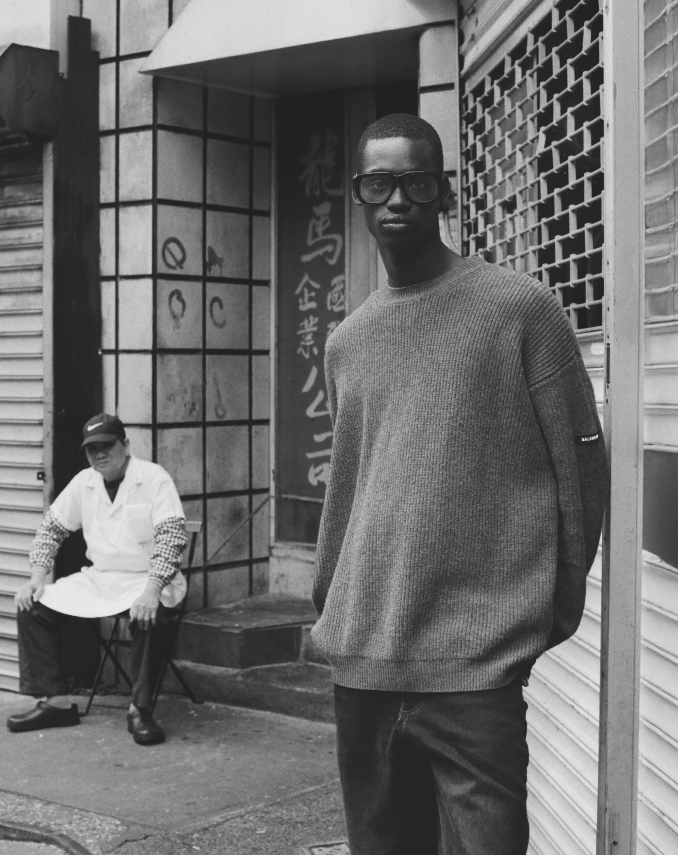 An adult man wearing a sweater and sunglasses poses in an urban environment near a seated man wearing a lab coat in black and white.
