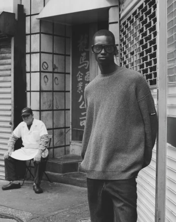 An adult man wearing a sweater and sunglasses poses in an urban environment near a seated man wearing a lab coat in black and white.
