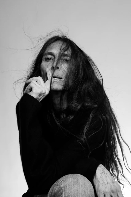Black and white portrait of a mature woman with long dark hair holding a finger to her lips, asking for silence in a light studio.