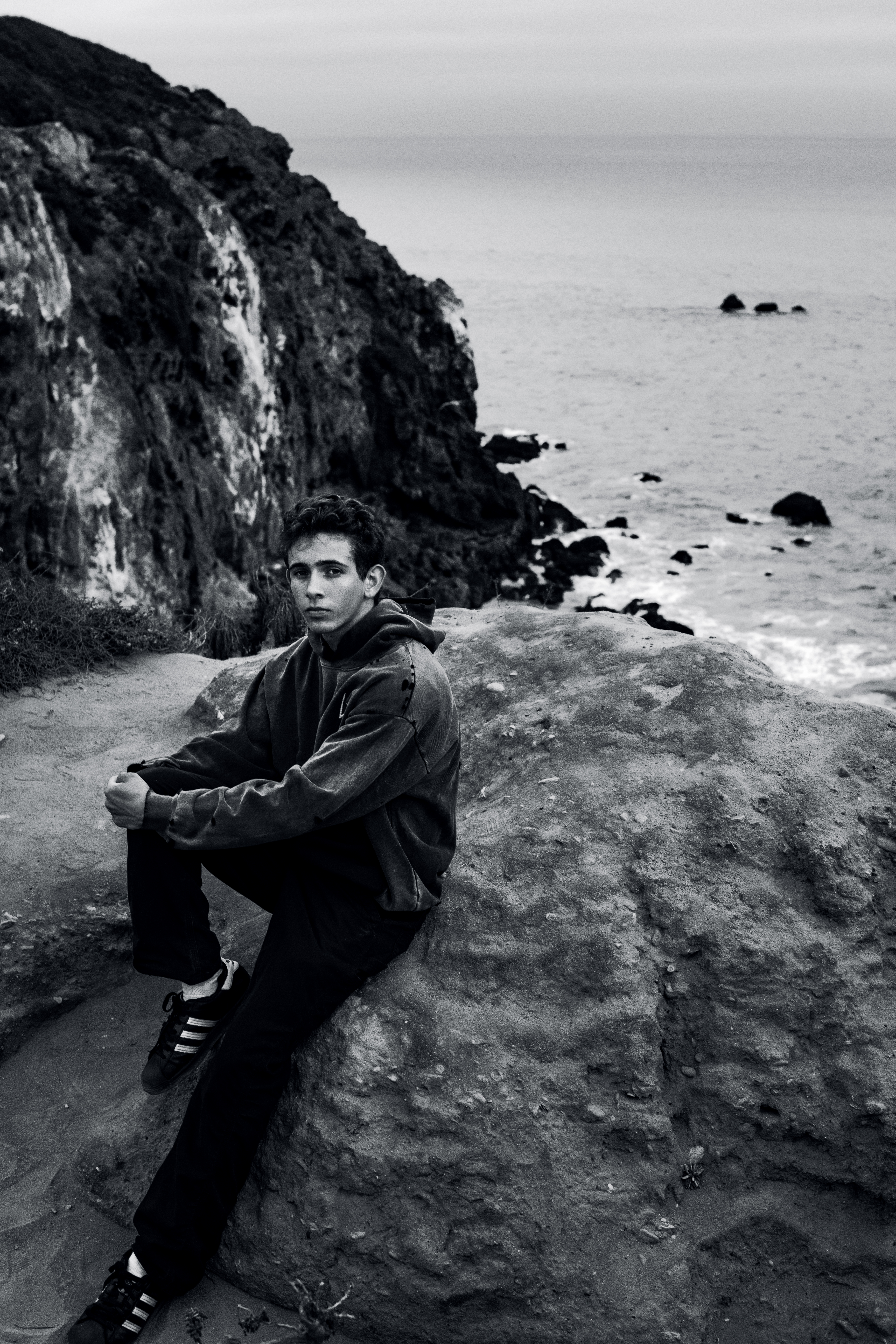 Young man in sweatshirt sitting on a rock looking at the ocean, with a cliff in the background, in a black and white photograph.