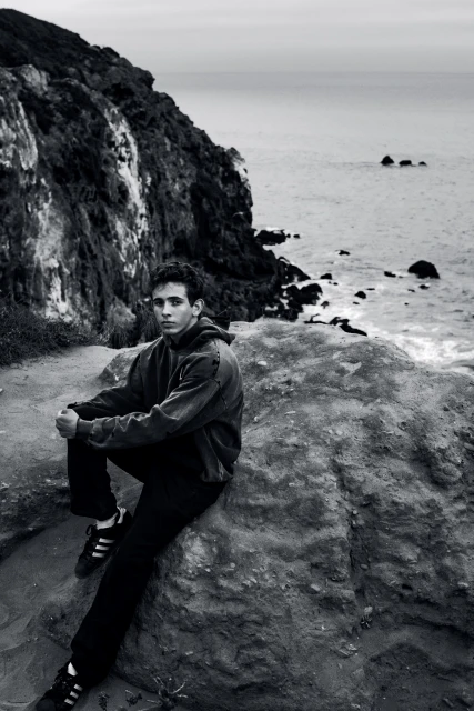 Young man in sweatshirt sitting on a rock looking at the ocean, with a cliff in the background, in a black and white photograph.