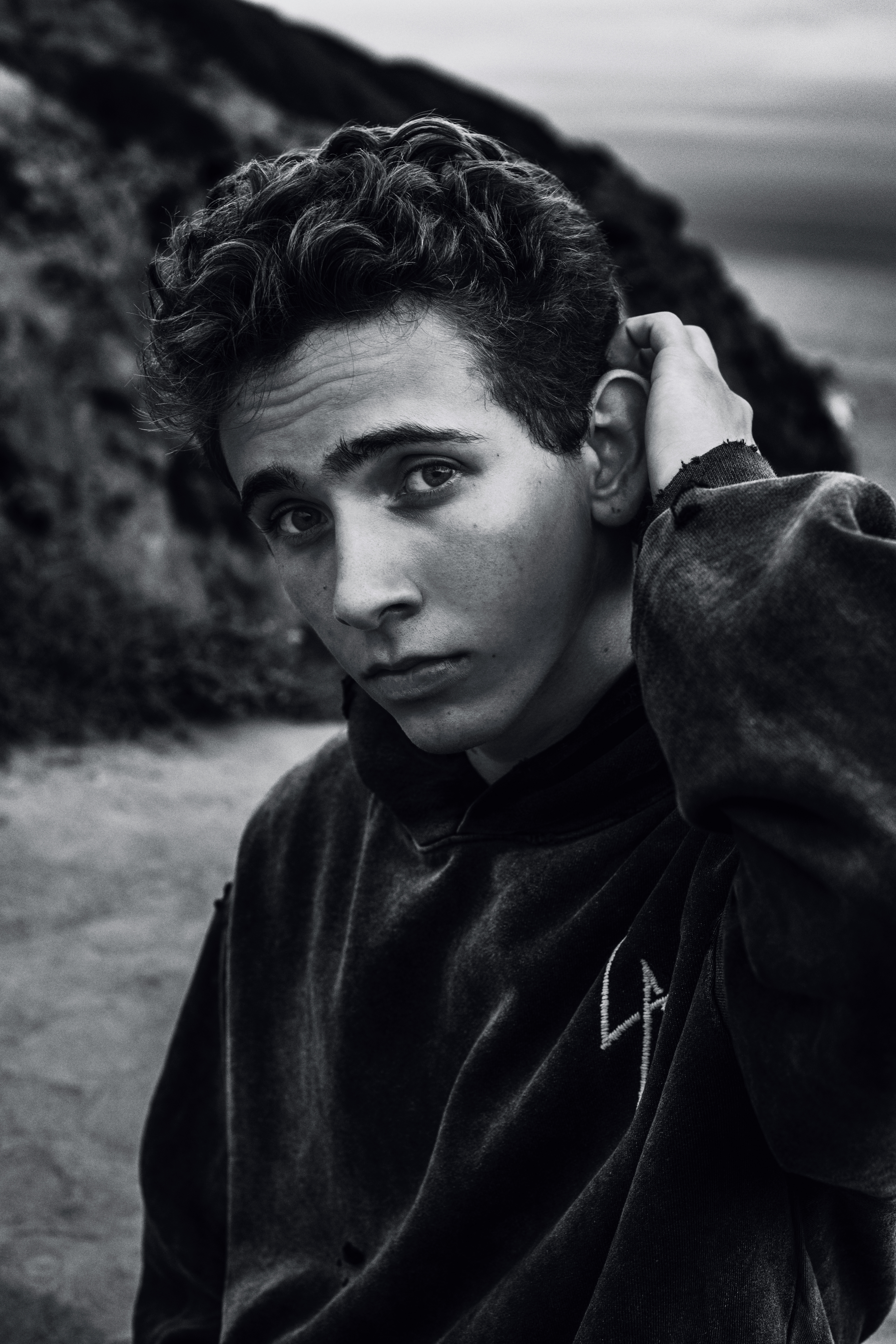 Monochrome portrait of a young adult with curly dark hair wearing a hoodie, outdoors in front of a blurred background.