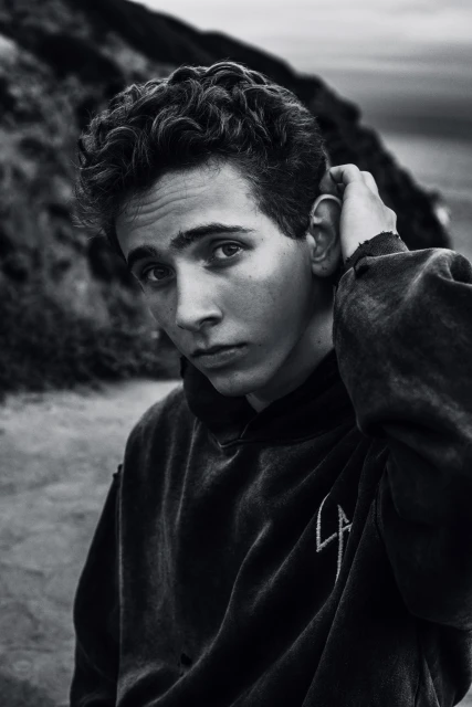 Monochrome portrait of a young adult with curly dark hair wearing a hoodie, outdoors in front of a blurred background.