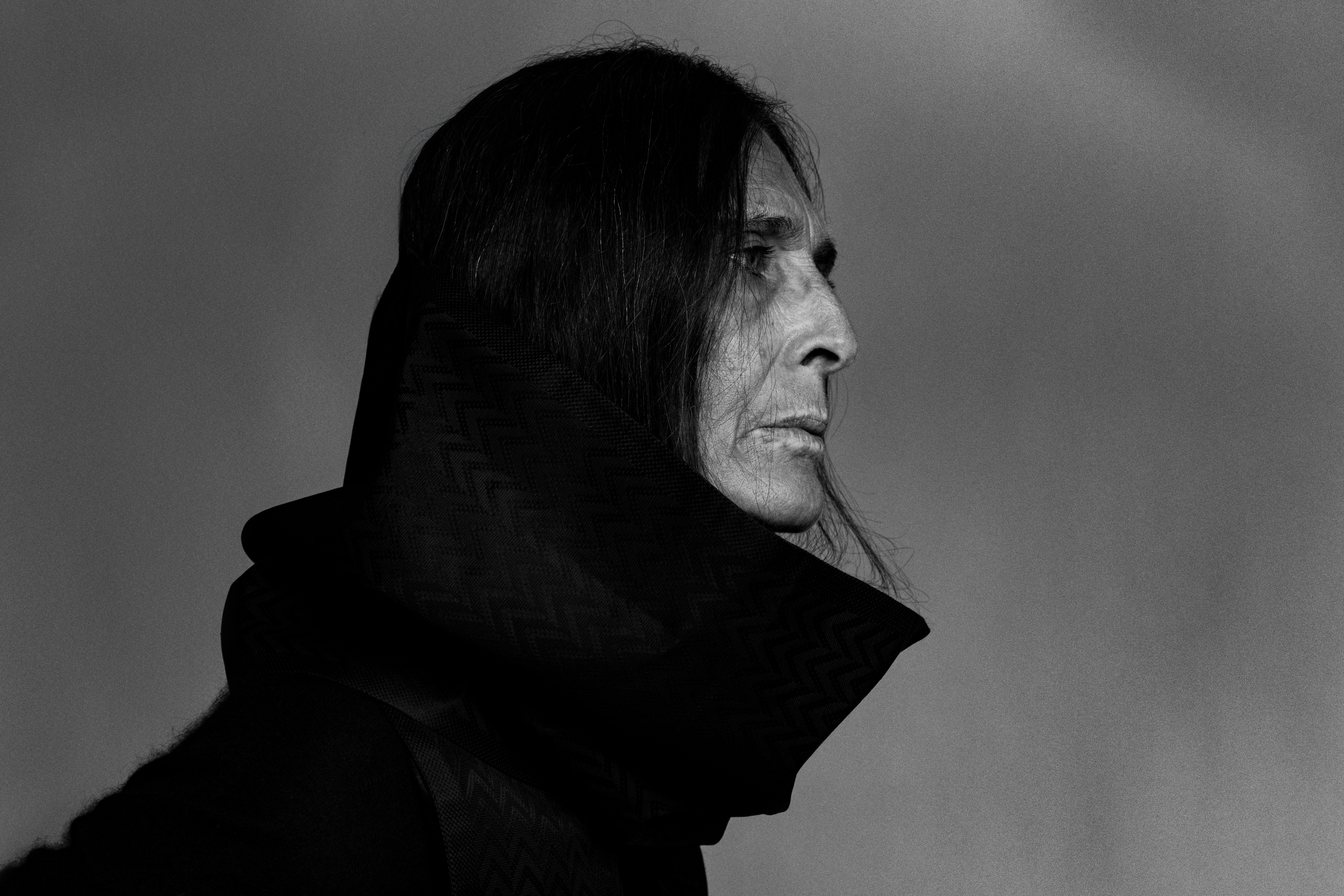 Monochrome portrait of an adult with long dark hair wearing a textured black garment, gazing to the right against a gray background.