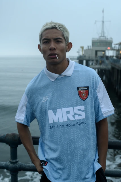 Young adult in light blue Martine Rose soccer jersey stands near the ocean on a pier with gray skies and vintage style processing.