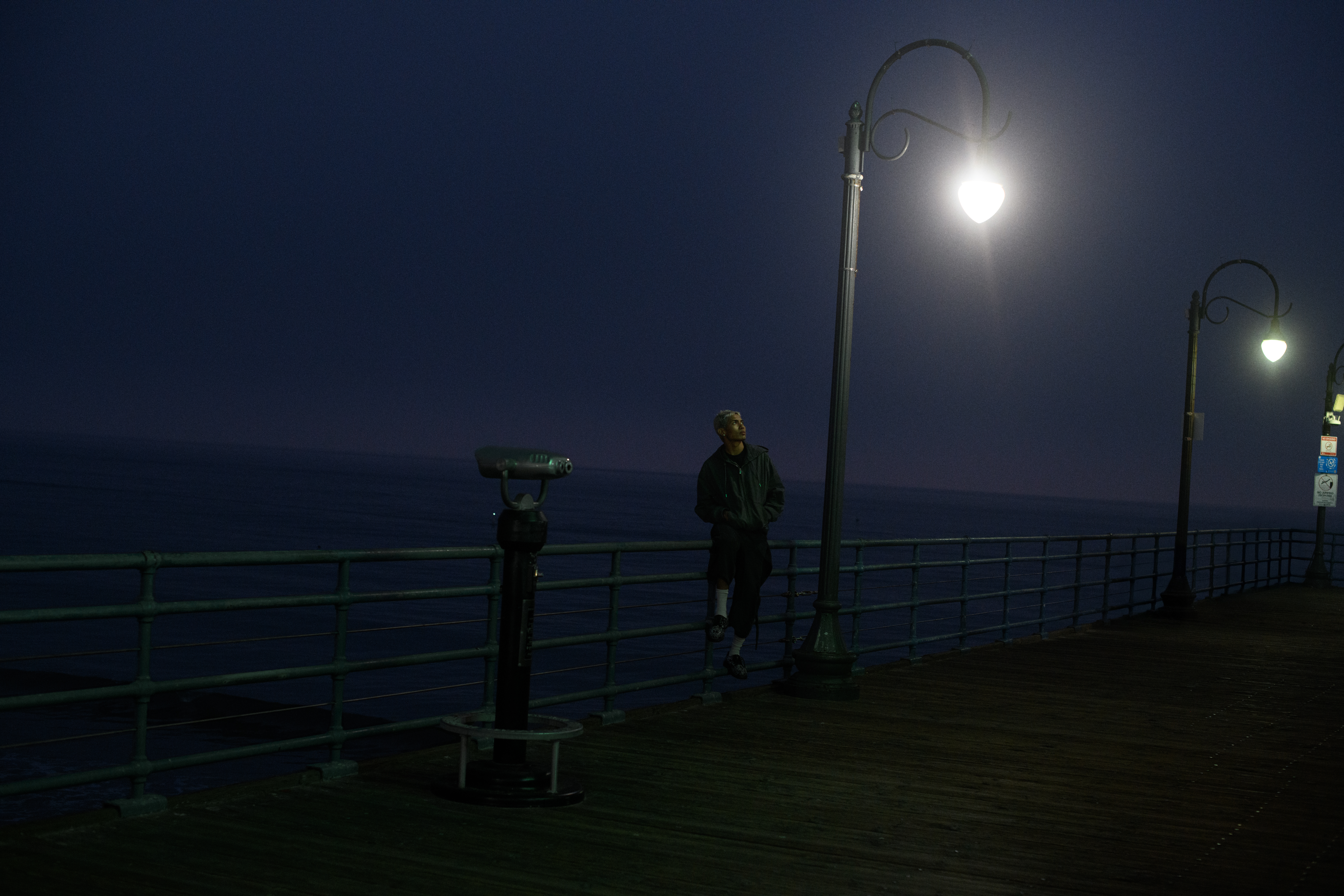 Man stands on a pier at night under a lamppost near a telescope. Dark water and sky create a moody, atmospheric scene.