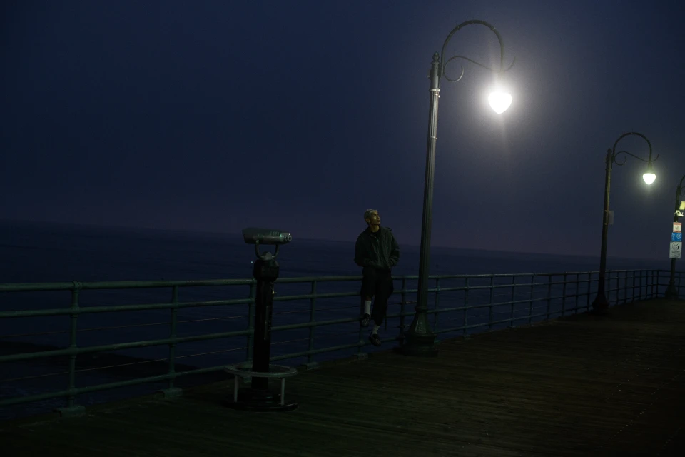 Man stands on a pier at night under a lamppost near a telescope. Dark water and sky create a moody, atmospheric scene.
