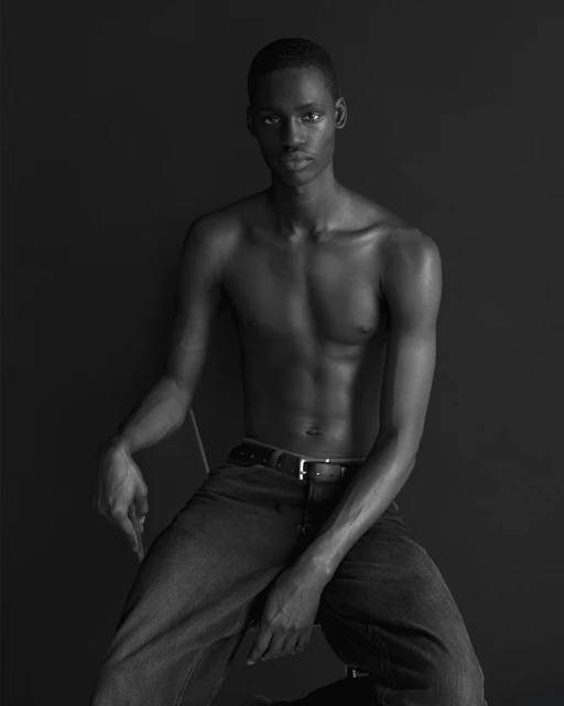 Black and white studio shot of a shirtless African American man with jeans, seated against a dark background, with a neutral facial expression.
