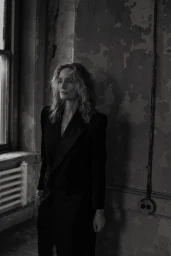 Monochrome portrait of a woman with wavy hair wearing a tailored dark blazer standing near a window.