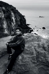 Young man in a hoodie and sneakers sitting on a rocky cliff overlooking the ocean in black and white.