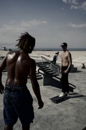 Two shirtless skateboarders at a concrete skate park next to a sandy beach in Los Angeles.