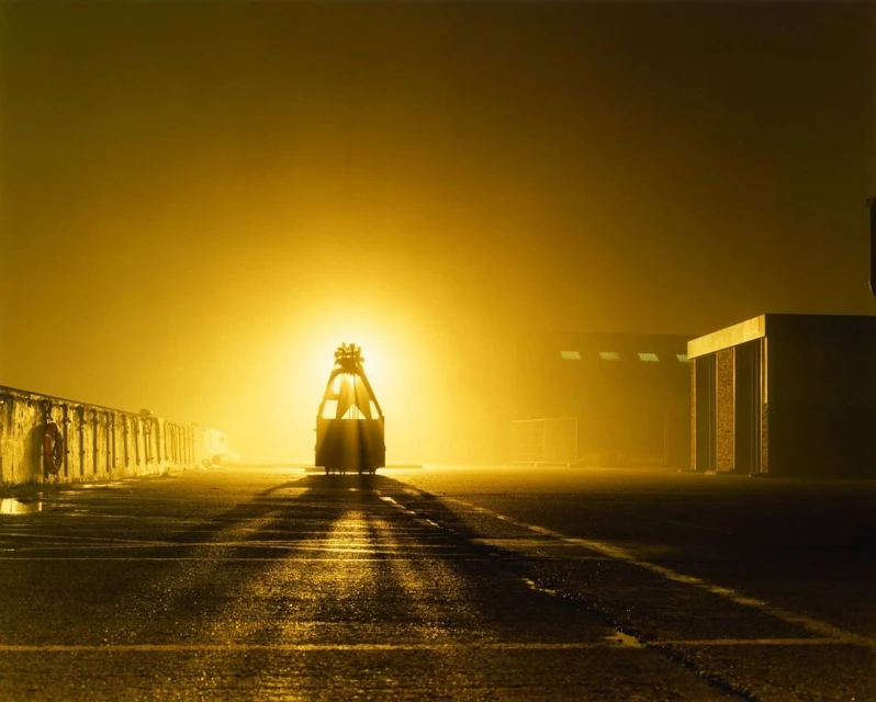 Fog-shrouded flatbed car on tracks in a dark outdoor setting with yellow lighting and industrial buildings in the distance.