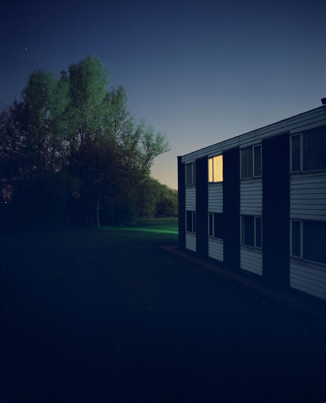 Apartment building exterior at night with glowing windows, framed by green trees under a dark blue sky.