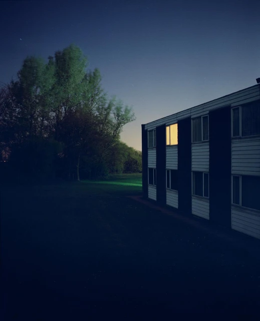 Apartment building exterior at night with glowing windows, framed by green trees under a dark blue sky.