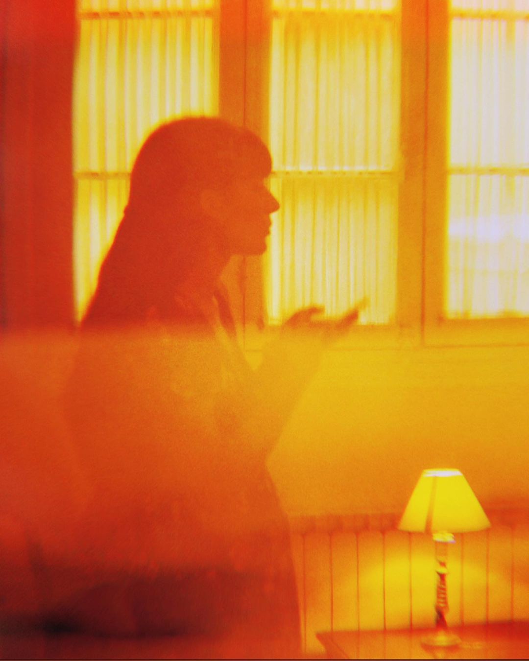 Silhouette of a woman with dark hair in front of a window with curtains, next to a lamp on a table, interior setting with warm lighting.